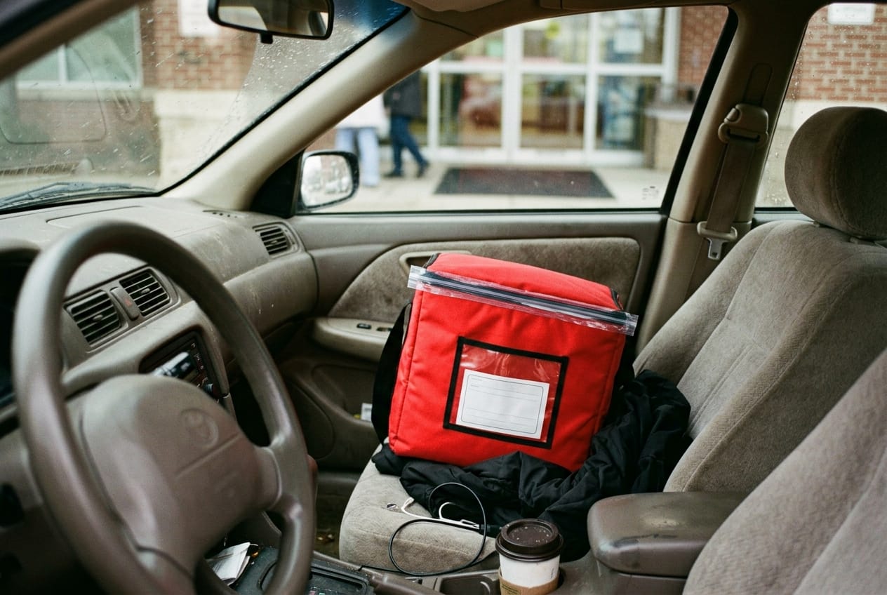 Sealed red STAT delivery bag on the passenger seat of a courier car, hospital pharmacy entrance blurred through the windshield